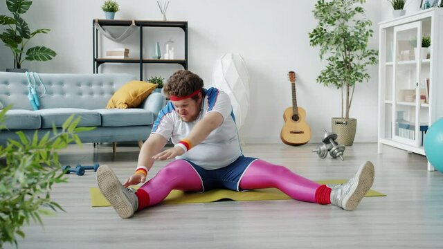 Awkward overweight young man in bright outfit is stretching legs and arms then falling on floor feeling tired exercising alone at home. People and sports concept.
