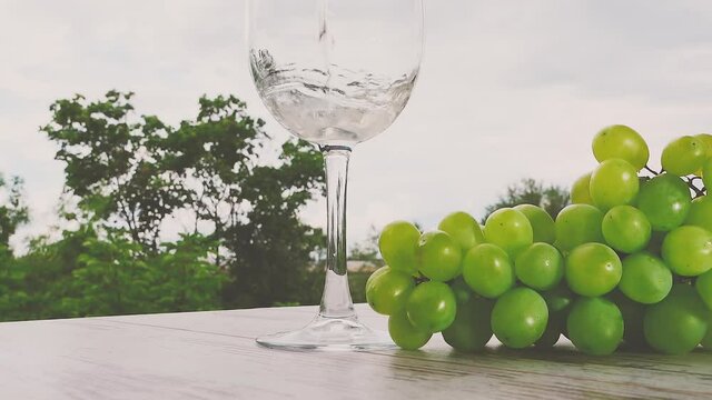 A hand pours white wine from a bottle into a glass that stands on a wooden table with a branch of green grapes against the background of nature.