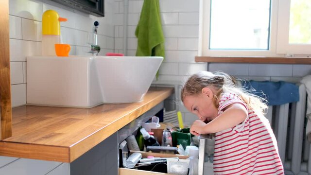 Fair haired preschooler girl in striped t-shirt opens large kitchen drawer and looks inside with interest searching tools at home