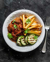 Slow cooker beef stew with grilled zucchini and french fries on a dark background, top view