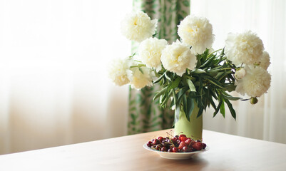 A bouquet of peonies in a vase and a plate with cherries. Summer concept