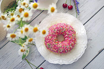 Donut in pink glaze on a background of wild flowers of chamomiles, fresh berries on a wooden table