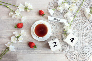 Calendar for July 24 : cubes with the number 24, the name of the month of July in English, a cup of tea on a gray openwork napkin, scattered flowers of bluebells and strawberries, top view