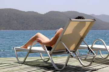 Woman sunbathing in lounge chair on wooden pier on sea and misty mountains background. Beach vacation, summer leisure and travel