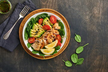 Fresh green salad with grilled chicken fillet, spinach, tomatoes, avocado, lemon and black sesame seeds in white bowl on old wooden dark table background. Nutrition Diet Concept. Top view .