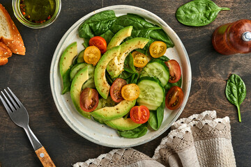 Avocado, red, yellow, black cherry tomato, spinach and cucumber fresh salad with spices pepper and olive oil in grey bowl old wooden table background. Healthy food concept. Top view.
