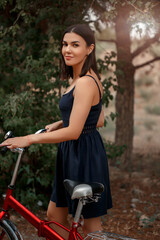 Fototapeta na wymiar a young brunette woman of Asian appearance in a dark blue dress is standing next to a bicycle with a punctured wheel. there is a grain and a small depth of focus in the photo for artistic purposes