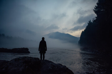 Alone human silhouette in white haze against the mountains and river. Thick fog in the evening...