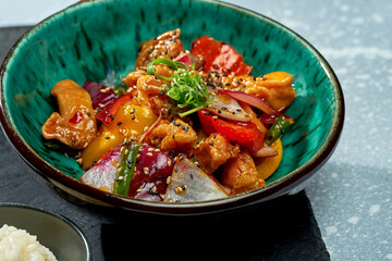 Appetizing pan-Asian dish - chicken wok in sweet and sour sauce with bell pepper, onion, sesame seeds and rice garnish in a blue bowl on a gray background