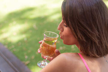 Beautiful woman in bright pink top wears beaded necklace sits on grass in summer outdoor cafe smiling holds fresh cool drink juice lemonade