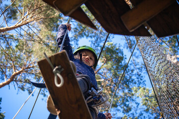 Obraz na płótnie Canvas a girl in a green helmet with a safety net, smiling, runs along wooden hanging boards a distance in a forest extreme rope park