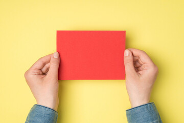 First person top view photo of female hands holding red paper card on isolated yellow background with blank space