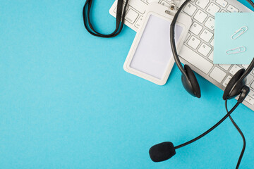 Top view photo of sticker note clips id card holder and black headphones with microphone on white keyboard on isolated pastel blue background with blank space