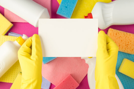 Top view photo of hands in yellow rubber gloves holding white paper card detergent bottles multicolor rags sponges on isolated pink background with copyspace