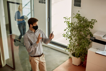 Happy businessman with face mask waving during video call via smart phone at work.