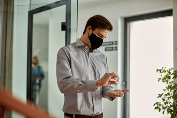 Naklejka na ściany i meble Male entrepreneur with face mask disinfecting his hands in hallway due to COVID-19 pandemic.