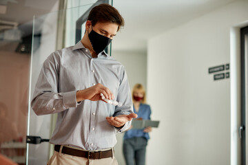 Businessman with face mask disinfecting his hands while working in the office during coronavirus pandemic.