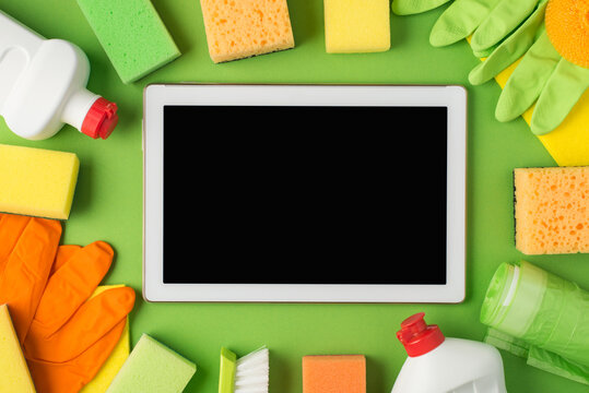 Top view photo of tablet computer in the middle detergent bottles rubber gloves rags sponges brush and garbage bags on isolated green background with copyspace