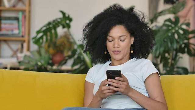 Portrait of a young African American woman texting on her phone and enjoys good news. Brunette with curly hair sitting on yellow sofa in a bright home room. Close up. Slow motion.