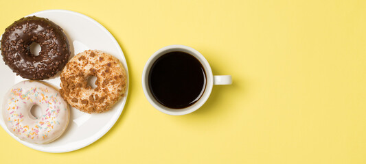 Top view photo of cup of coffee and plate with three different donuts on isolated light yellow background with empty space