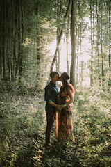 Naklejka na ściany i meble Happy pregnant woman and her man, standing hugging each other, posing on the background of beautiful sunset in the forest among trees, and kissing. Happy pregnancy concept