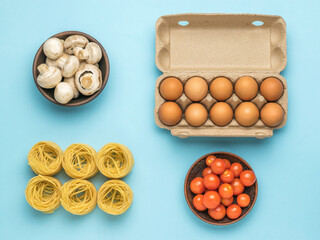 A bowl of tomatoes, a bowl of mushrooms, pasta and a box of eggs on a blue background.