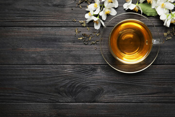 Glass cup of aromatic jasmine tea, dry leaves and fresh flowers on black wooden table, flat lay. Space for text