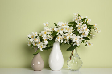 Beautiful jasmine flowers in vases on table near light green wall
