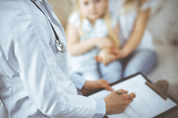 Fototapeta premium Doctor and patient. Pediatrician using clipboard while examining little girl with her mother at home. Sick and unhappy child at medical exam. Medicine concept