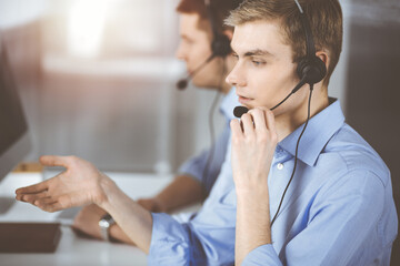 Two young men in headset, sitting at the desk in a sunny modern office, listening to the clients. Call center operators at work