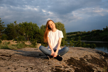 Naklejka na ściany i meble Nature, wellbeing, Relaxation, meditation and mental health. Interacting, Thriving with nature. Calming and enchanting effect. Young redhead woman sitting on rock and watching on river on sunset