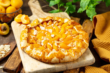 French Galette or a pie with apricots and almonds on on a wooden serving board on a brown culinary background closeup. Summer homemade sweet pastries