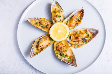Baked Greenshell mussels in half a shell with cheese, parsley and lemon on a white plate. Traditional hot appetizer. Close-up, top view.