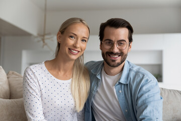 Head shot portrait of smiling couple making video call together, sitting on couch at home, happy young woman and man in glasses looking at camera, chatting with friends online, using webcam