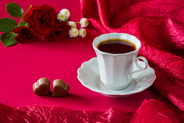 Coffee in a white cup, two heart-shaped candies and rose flowers. Romantic concept in dark red tones