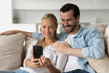 Smiling young couple hugging using smartphone sitting on cozy couch together, happy woman and man in glasses looking at phone screen, watching video, chatting, satisfied clients shopping online