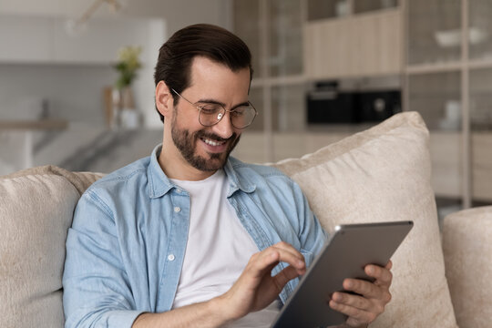 Close up smiling man in glasses using tablet at home, sitting on cozy couch, happy young male looking at modern device screen, typing writing message in social network having fun browsing apps