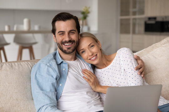 Head shot portrait of happy romantic couple hugging, using laptop together, smiling wife and husband looking at camera, sitting relaxing on couch at home, enjoying leisure time with device