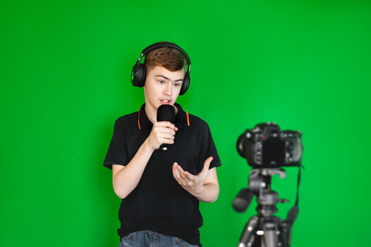 A boy teen blogger shoots a video on camera against a green background. A teenage Caucasian in a black T-shirt emotionally tells something for a blog.