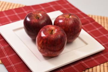 Three red apples on a square plate with a red checkered tablecloth