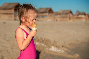 Cute little girl in a pink swimsuit with ice cream in a cone in hand on the sandy beach by the sea