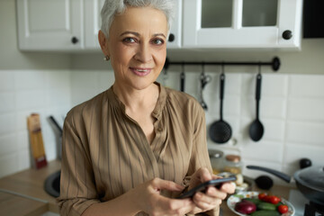 Cheerful woman holding smartphone in hands. Smiling middle-aged housewife posing in kitchen and looking at camera with happiness. Household and housekeeping concept