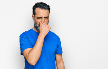 Middle aged man with beard wearing casual blue t shirt smelling something stinky and disgusting, intolerable smell, holding breath with fingers on nose. bad smell