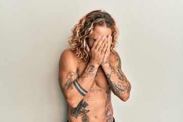 Handsome man with beard and long hair standing shirtless showing tattoos with sad expression...