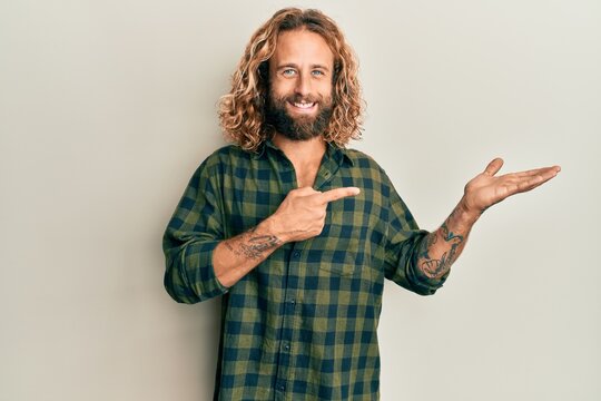 Handsome man with beard and long hair wearing casual clothes amazed and smiling to the camera while presenting with hand and pointing with finger.