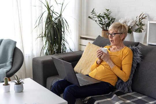 An older woman with short blonde hair sits on sofa in living room and chats with friend on laptop over video chat