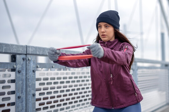 Focused young fitness woman in winter sportswear, winter skully hat and winter gloves work out with elastic resistance band in hands on bridge during cold day