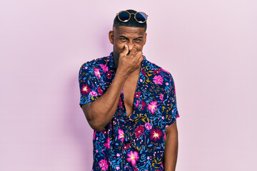 Young black man wearing hawaiian shirt and sunglasses smelling something stinky and disgusting, intolerable smell, holding breath with fingers on nose. bad smell