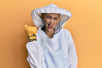 Beautiful blonde caucasian woman wearing protective beekeeper uniform angry and mad raising fist...