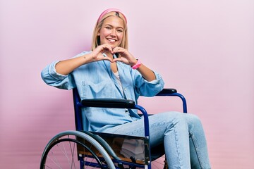 Beautiful blonde woman sitting on wheelchair smiling in love doing heart symbol shape with hands....
